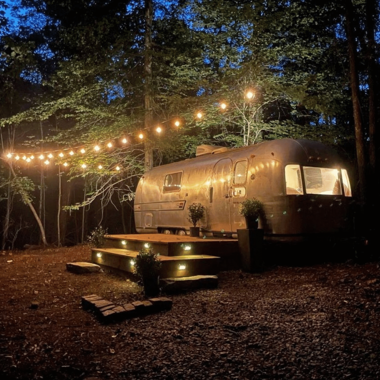 Caravane Airstream nuit guirlandes lumineuses terrasse éclairée forêt ambiance cosy